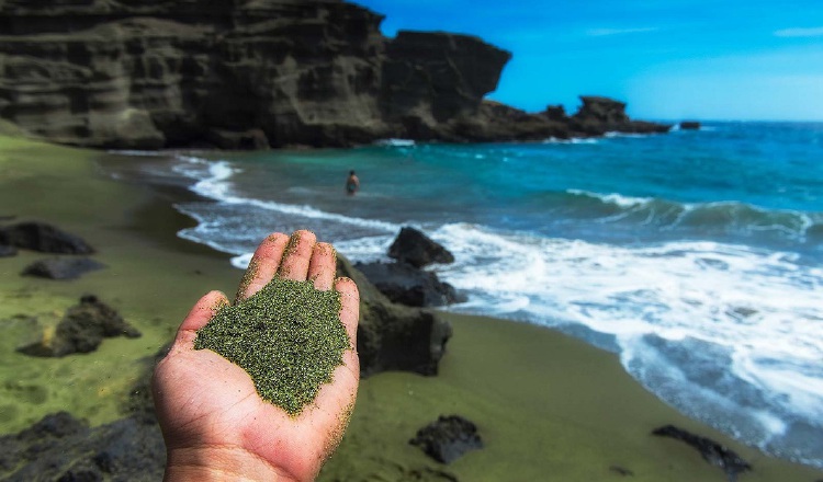 Mano con olivina molida en primer plano y al fondo una playa con arenas verdes. Project Vesta y Climitigation